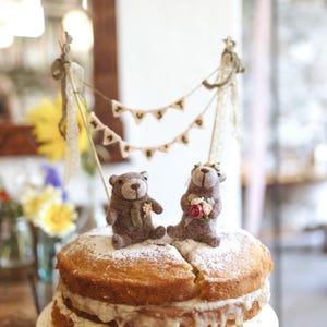 May include: A two-layer sponge cake with cream filling and powdered sugar topping, featuring two brown felt bear figurines. The bears are positioned under a miniature bunting banner with the text JUST MARRIED, suspended between two wooden sticks.