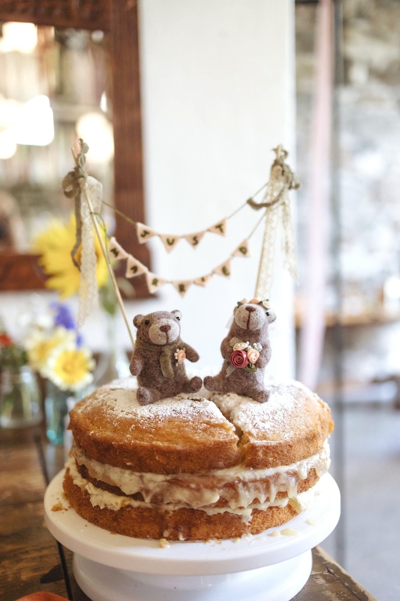 May include: A two-layer sponge cake with cream filling and powdered sugar topping, featuring two brown felt bear figurines. The bears are positioned under a miniature bunting banner with the text JUST MARRIED, suspended between two wooden sticks.