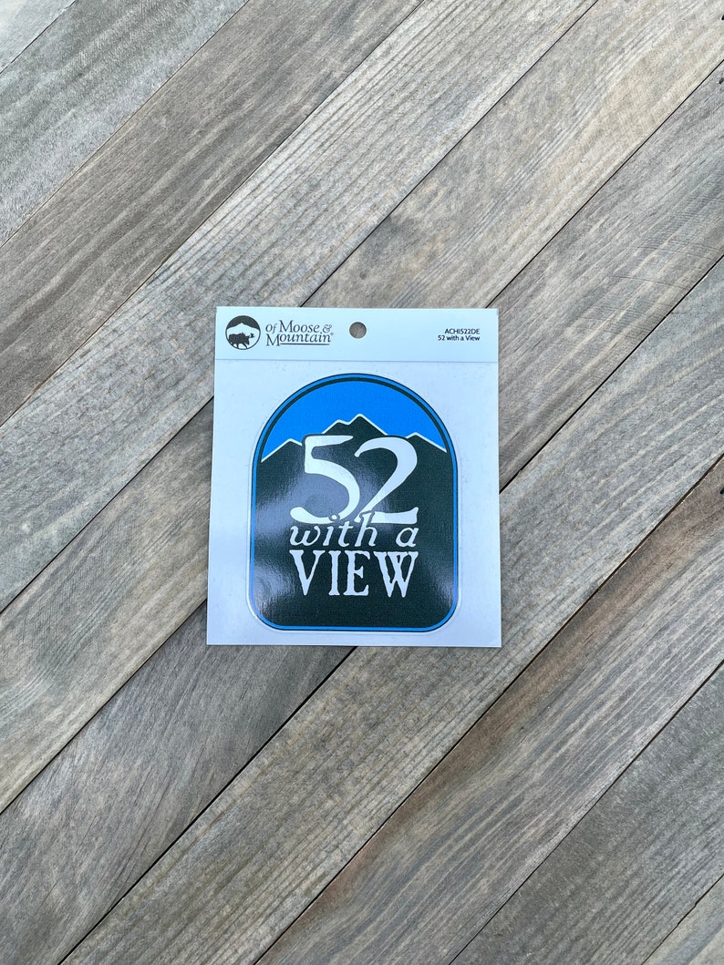 May include: A blue and green sticker with the text "52 with a VIEW" and a mountain range graphic. The sticker is from "of Moose & Mountain" and is an "ADHESIVE STICKER".