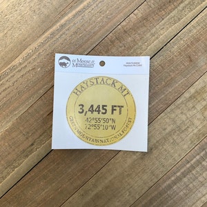 May include: A sticker featuring a circular design with the text "HAYSTACK MT 3,445 FT" in a vintage style. The sticker also includes the coordinates 42°55'50"N 72°55'10"W and the words "GREEN MOUNTAIN NATIONAL FOREST". The sticker is on a white background.