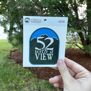 May include: A white sticker with a blue and green circle design. The circle has a white number "52" with the text "with a VIEW" below it. The sticker is from "of Moose & Mountain" and has the product code "ACH152DE".