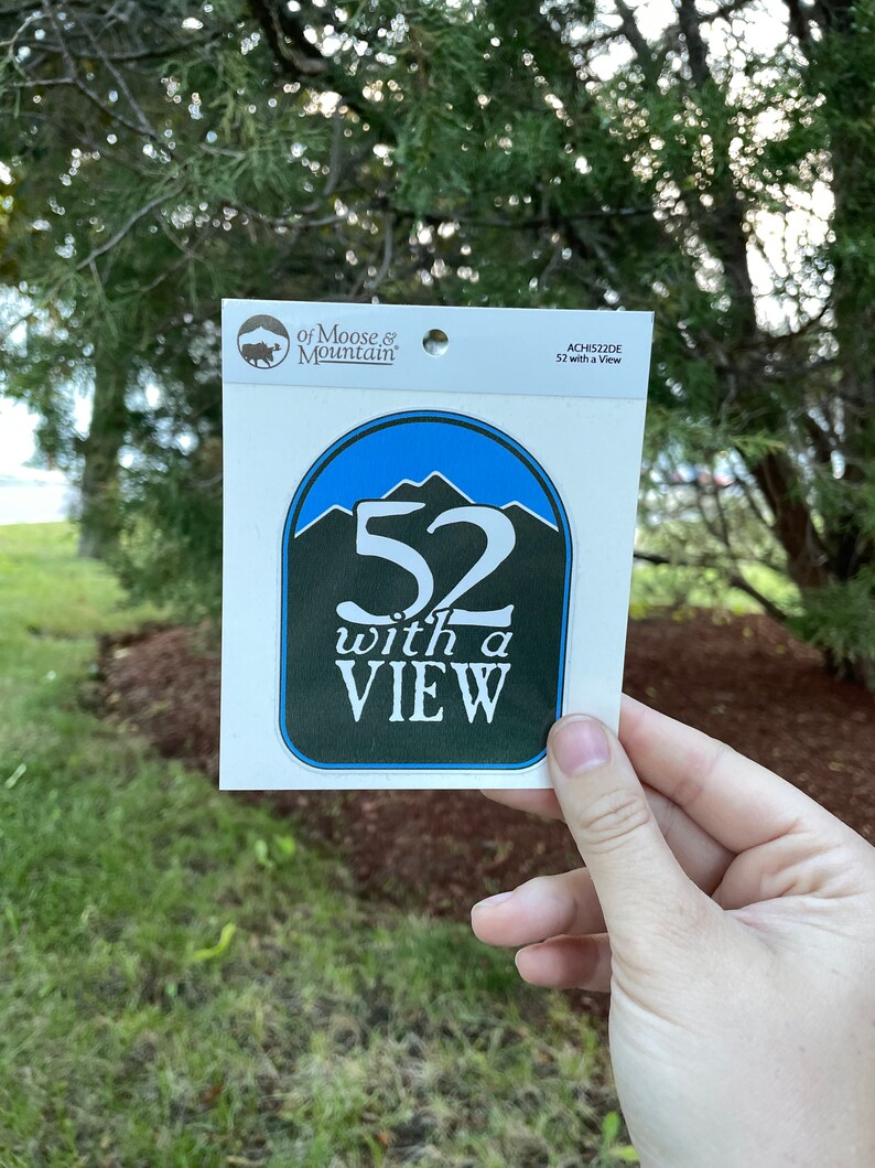 May include: A white sticker with a blue and green circle design. The circle has a white number "52" with the text "with a VIEW" below it. The sticker is from "of Moose & Mountain" and has the product code "ACH152DE".