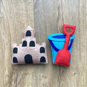 May include: A felt toy set featuring a light brown castle with black arched windows and a blue bucket with a red shovel. The toys are placed on a wooden surface.