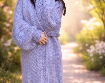 May include: A long, light purple knit cardigan with a relaxed fit. The cardigan features long sleeves and a floor-length design. The soft, textured knitwear is a solid color, creating a cozy and elegant look. The model is wearing the cardigan.