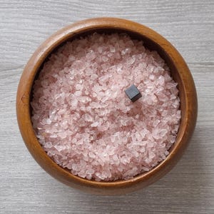 Pode incluir: Uma tigela redonda de madeira cheia de pequenos fragmentos de cristal de quartzo rosa translúcido. Um único cubo pequeno e sólido de cor preta está posicionado perto do centro do monte de pedras cor-de-rosa, sobre os fragmentos de cristal.