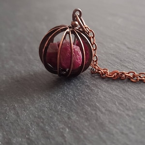 Könnte beinhalten: Eine Kupferkette mit einem Käfiganhänger, der kleine rote Rubin-Steine enthält.
