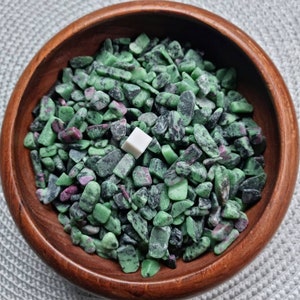May include: A bowl filled with green and black tumbled stones. The stones are small and have a rough texture. There is a small white square in the centre of the bowl.