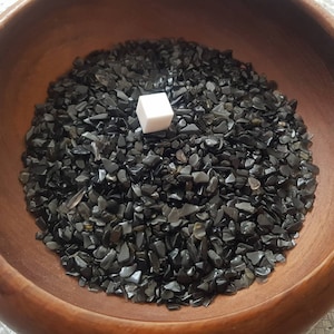 Könnte beinhalten: Eine Holzschale gefüllt mit schwarzen Obsidian-Chips. Ein kleiner weißer Würfel befindet sich in der Mitte der Schale.