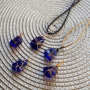 May include: Four necklaces with blue glass pendants wrapped in gold wire. The pendants are different shapes and sizes. The necklaces are on a woven placemat.