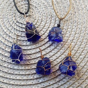 May include: Five blue glass pendants wrapped in wire, each with a different wire color. The pendants are hanging from cords on a woven background.