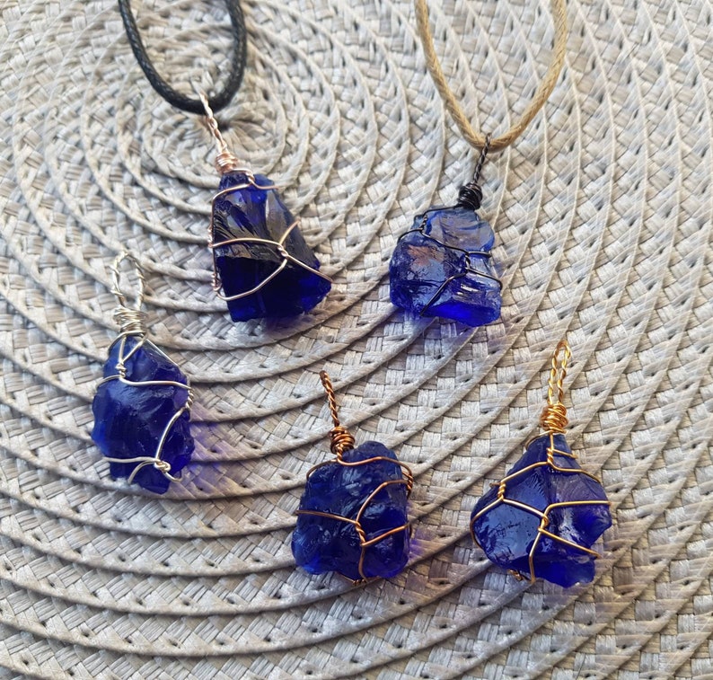May include: Five blue glass pendants wrapped in wire, each with a different wire color. The pendants are hanging from cords on a woven background.