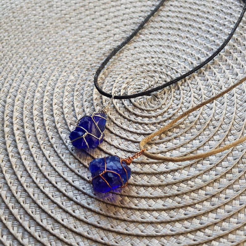 May include: Two necklaces with blue glass pendants wrapped in wire. One necklace has a black cord and the other has a brown cord.
