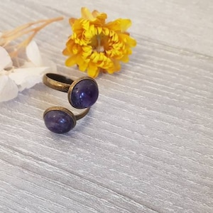 May include: A bronze ring with two purple gemstones. The ring is on a white wooden surface with a yellow flower.