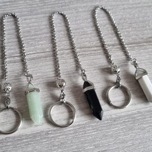 May include: Five silver chains with different coloured crystal pendants. The pendants are in the shape of a point and are attached to a silver ring. The crystals are green, black, and white.