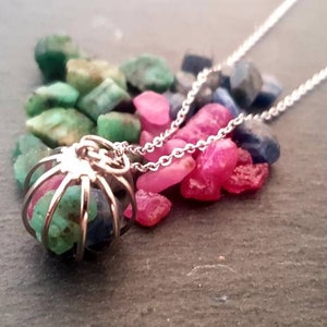 Könnte beinhalten: Eine silberne Kettenkette mit einem Käfiganhänger, der eine Mischung aus grünen, rosa und blauen Edelsteinen enthält. Der Anhänger ist von losen Edelsteinen in denselben Farben umgeben.