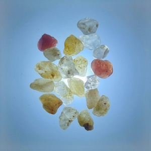 May include: A collection of small, rough, natural gemstones in various shades of pink, yellow, and white. The stones are arranged in a cluster on a light blue background.