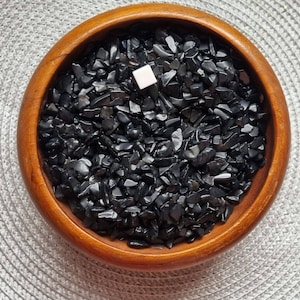 Pode incluir: Uma tigela de madeira cheia de lascas de obsidiana preta. A tigela está sobre uma superfície com um padrão cinza e branco.
