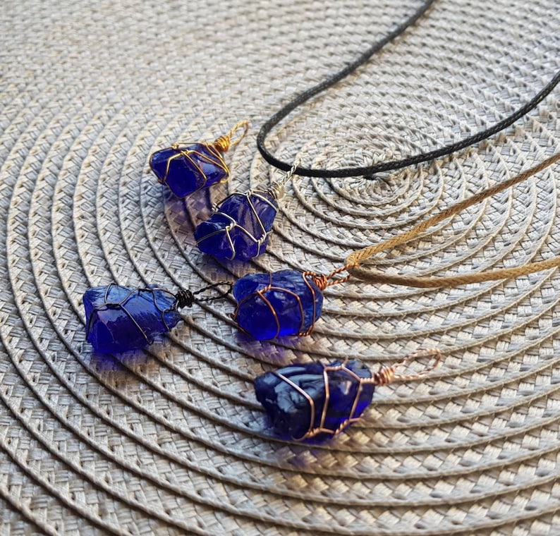 May include: Four cobalt blue sea glass pendants wrapped in copper wire, hanging from cords on a woven surface.