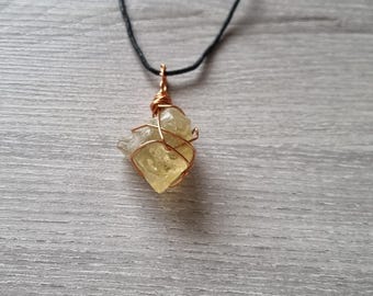 Puede incluir: Un colgante de cristal de citrino amarillo en bruto, envuelto en alambre de cobre, suspendido de un cordón negro. El colgante tiene una forma irregular natural, que resalta la textura y el color únicos de la piedra. El alambre de cobre añade un elemento artesanal rústico.
