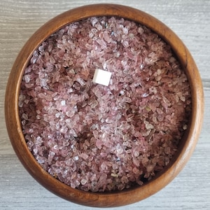 May include: A wooden bowl filled with pink and clear crystal chips. The bowl is on a light grey surface.
