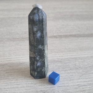 Könnte beinhalten: Eine dunkelgraue und weiße Kristallspitze mit einer spitzen Oberseite und einer quadratischen Basis. Ein kleiner blauer Würfel befindet sich vor der Kristallspitze.