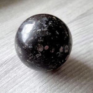 May include: A dark sphere with a polished finish, showing a mottled pattern of black, grey, and pinkish-white. The object sits on a light-coloured, textured surface. The sphere is a decorative item.