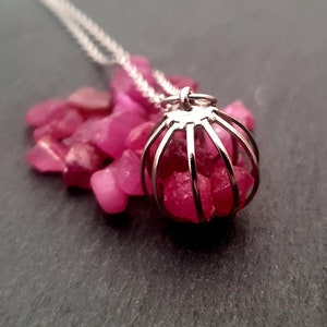 Könnte beinhalten: Eine silberne Käfig-Anhänger-Halskette mit einem Cluster aus rosafarbenen Rubin-Steinen. Die Halskette wird auf einer schwarzen Oberfläche mit lose verstreuten rosafarbenen Rubin-Steinen gezeigt.