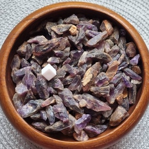 May include: A wooden bowl filled with amethyst crystal chips. The chips are various shades of purple and have a rough, natural texture.