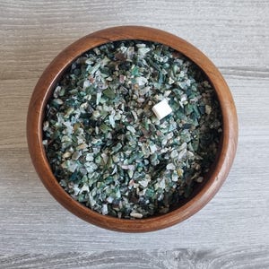 Puede incluir: Un cuenco de madera lleno de pequeñas piedras verdes y blancas de forma irregular. Un pequeño objeto cuadrado blanco descansa entre las piedras. El cuenco es de madera marrón claro y está sobre una superficie de madera gris claro.
