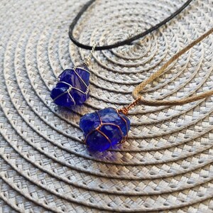 May include: Two necklaces with blue glass pendants wrapped in silver wire. One necklace has a brown cord and the other has a black cord.