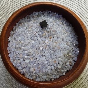 May include: A wooden bowl filled with light blue and white tumbled stones. The stones are small and smooth, and they are arranged in a random pattern. There is a small black square in the centre of the bowl.
