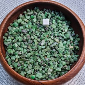 May include: A wooden bowl filled with small, green and black gemstones. The bowl is on a white and grey textured surface.