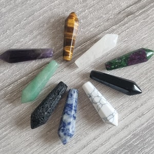 Op de afbeelding: Assortiment van gepolijste edelsteen toverstokken in verschillende kleuren en patronen, waaronder amethist, tijgeroog, jade en zwarte obsidiaan. Deze kristallen toverstokken zijn in een cirkelvormig patroon gerangschikt.