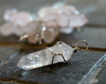 May include: A clear quartz crystal wrapped in silver wire, forming a pendant. The crystal is faceted and has a natural, raw look. The pendant is hanging on a gray surface.