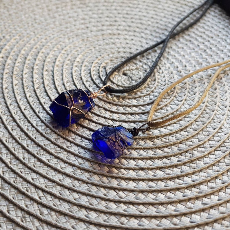 May include: Two necklaces with blue glass pendants wrapped in wire. The pendants are different shapes and sizes. The necklaces are on a light gray textured surface.