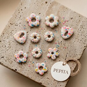 May include: A collection of ten small, white, flower-shaped and irregular-shaped charms with multicolored speckles in pink, blue, and yellow. A round white tag with the text PEPITA printed in black is attached with a thin brown twine loop.