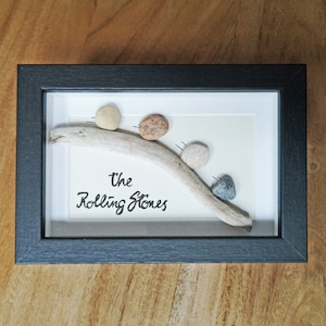 May include: A framed artwork with four small stones on a piece of driftwood. The stones are in shades of beige, brown, and grey. The words "The Rolling Stones" are written below the driftwood in black script.