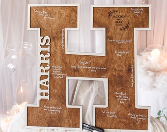 Puede incluir: Una letra H de madera con un marco blanco, de pie sobre un mantel blanco. La letra está decorada con un diseño floral y los nombres "Jackson" y "Ally" están escritos en ella. La letra está cubierta de escritura blanca, incluyendo buenos deseos para la pareja.