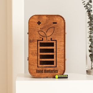Könnte beinhalten: Eine Holzkiste mit einem Batteriesymbol und den Worten "Gebrauchte Batterien" auf der Vorderseite. Eine einzelne AA-Batterie liegt auf einer weißen Oberfläche vor der Kiste.