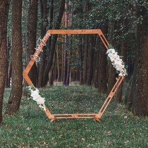 May include: A wooden hexagonal archway decorated with white flowers, standing in a woodland setting. The archway has the names "Emily & James" written on it.