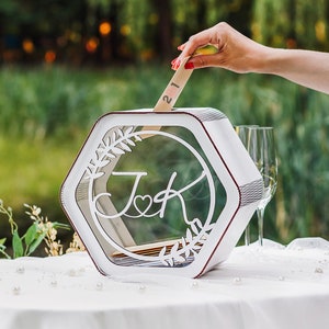 May include: White hexagonal wedding card box with a clear glass insert. The box has a cutout design of leaves and the initials "J & K" with a heart. A hand is placing a card with the number "21" into the box.