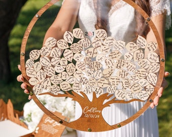 May include: A wooden tree-shaped guest book with a clear acrylic cover. The tree has a brown wooden trunk and branches with small wooden leaves that can be written on. The leaves are filled with handwritten messages. The tree is labeled "Collins 12/12/24".