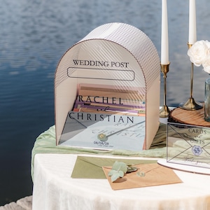 May include: A wedding card post with the words "Wedding Post" at the top. The names "Rachel and Christian" are printed on the front. Two white candles, a vase of flowers, and a "Cards & Gifts" sign are on a table.