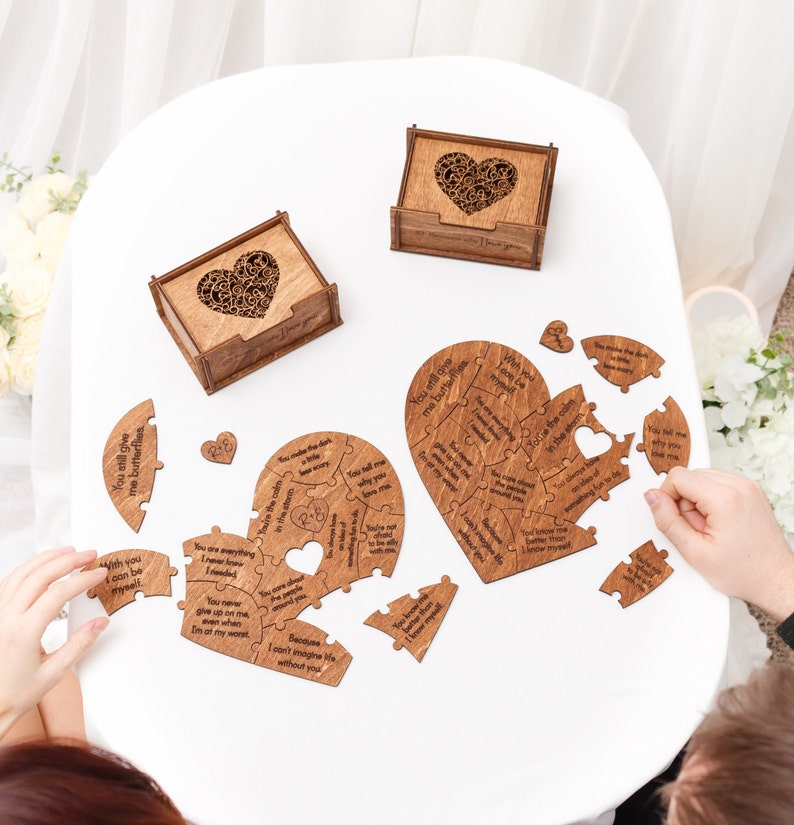 Puede incluir: Un rompecabezas de madera en forma de coraz&oacute;n con mensajes grabados para una pareja. El rompecabezas est&aacute; hecho de dos piezas que encajan para formar un coraz&oacute;n completo. Las piezas est&aacute;n esparcidas sobre una mesa blanca con dos peque&ntilde;as cajas de madera con recortes en forma de coraz&oacute;n.