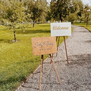 May include: Two wooden signs on easels. The top sign is white with gold lettering that says "Welcome Emily & James". The bottom sign is brown with white lettering that says "Emily & James June 24, 2023".
