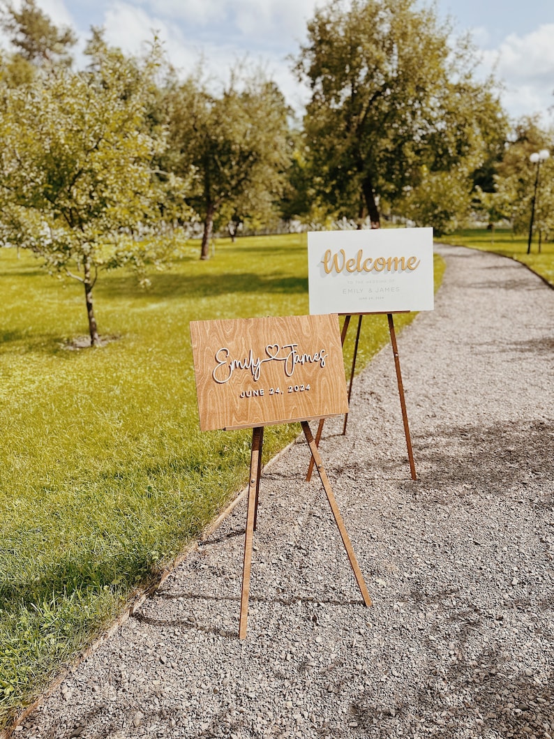 May include: Two wooden signs on easels. The top sign is white with gold lettering that says "Welcome Emily & James". The bottom sign is brown with white lettering that says "Emily & James June 24, 2023".