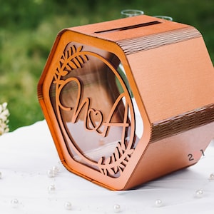 May include: A hexagonal wooden box with a clear acrylic window, decorated with a leafy design and the initials "N & A". The box is a light brown color and has a slot on the top for inserting cards or envelopes.