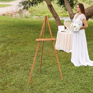 May include: A wooden easel stands on a grassy lawn. A white table with a white tablecloth is set up behind the easel. A bride in a white dress stands behind the table, holding a bouquet of white flowers.