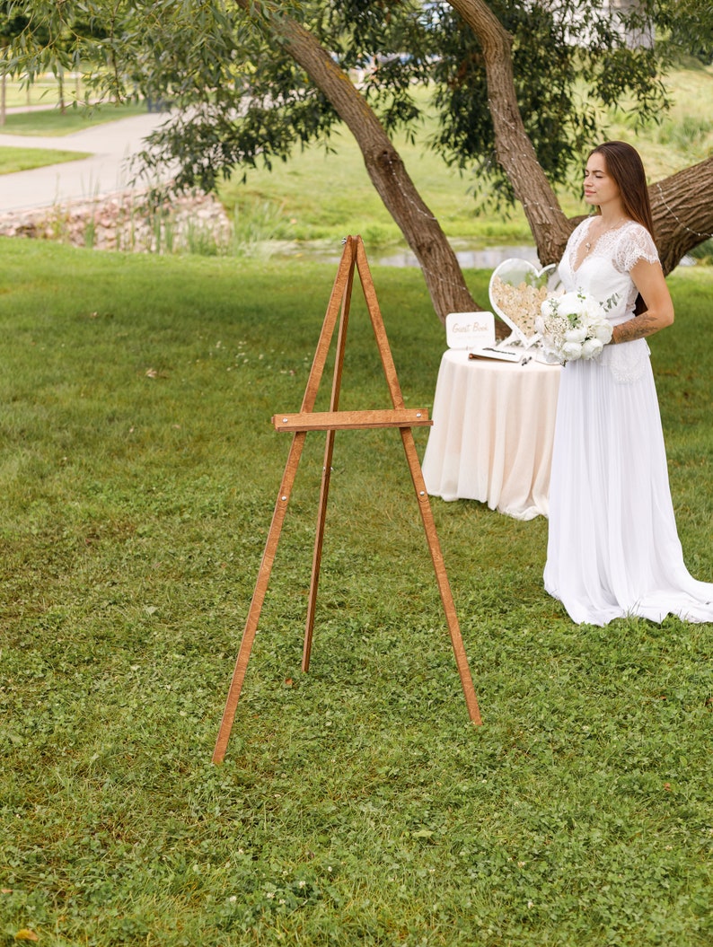 May include: A wooden easel stands on a grassy lawn. A white table with a white tablecloth is set up behind the easel. A bride in a white dress stands behind the table, holding a bouquet of white flowers.