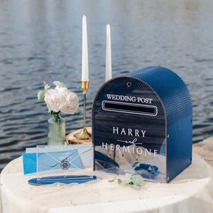 May include: A navy blue wedding card box with the words "Wedding Post" and the names "Harry and Hermione" in white script. The box is on a table with flowers and candles.